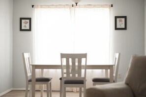 A dining area with a table and chairs, a window with sheer curtains, and two framed pictures on the wall.