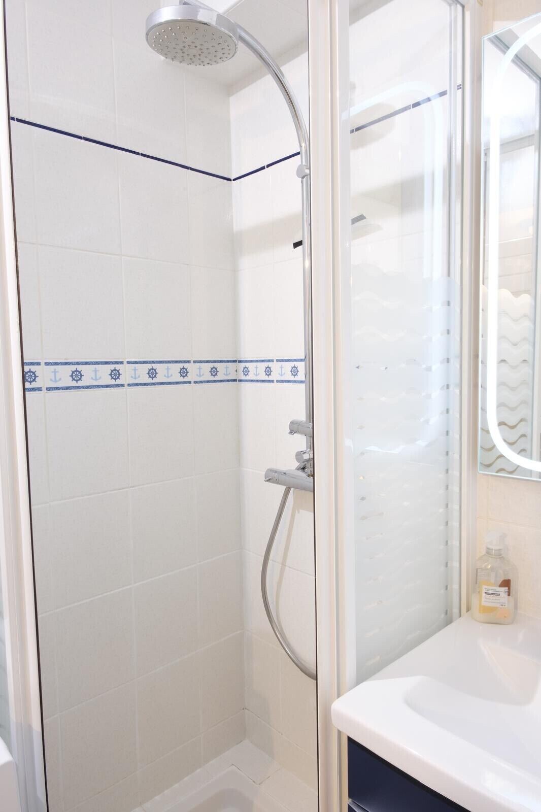 A shower with a glass door, a white tiled wall with blue decorative tiles, and a white sink with a mirror above it.