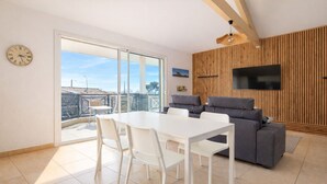 A modern living room with a dining table, chairs, a grey sofa, a flat-screen TV, and a wooden wall panel. - Charming apartment with terrace in Arès (Arès)