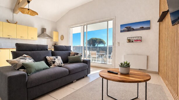 A modern living room with a grey sofa, a round wooden coffee table, and a large sliding glass door leading to an outdoor patio. - Charming apartment with terrace in Arès (Arès)