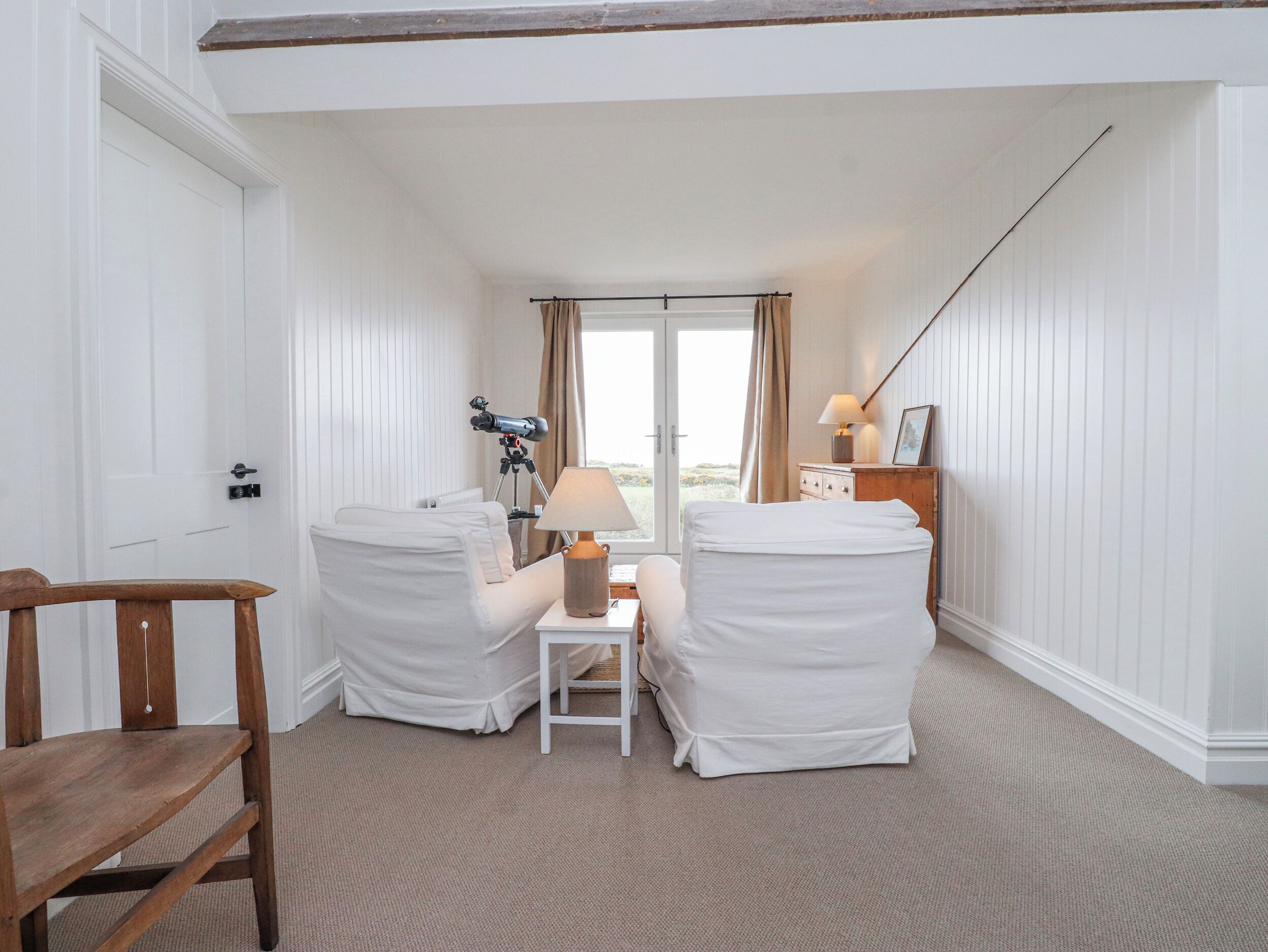 A room with two white-covered chairs, a small table with a lamp, a telescope, and a wooden chair.