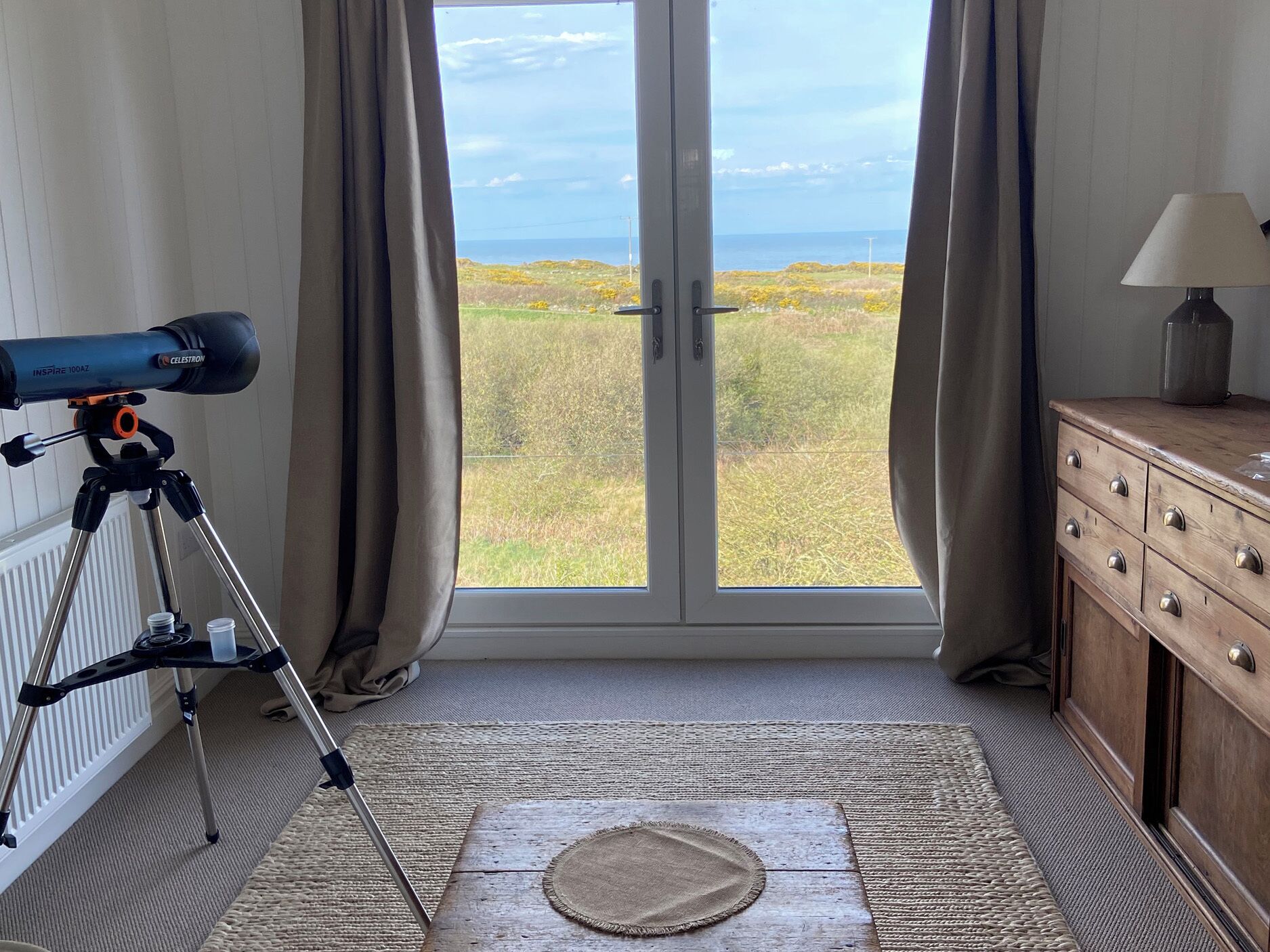 Una habitación con un telescopio sobre un trípode, vista al mar a través de un ventanal de vidrio, cómoda de madera y una lámpara.