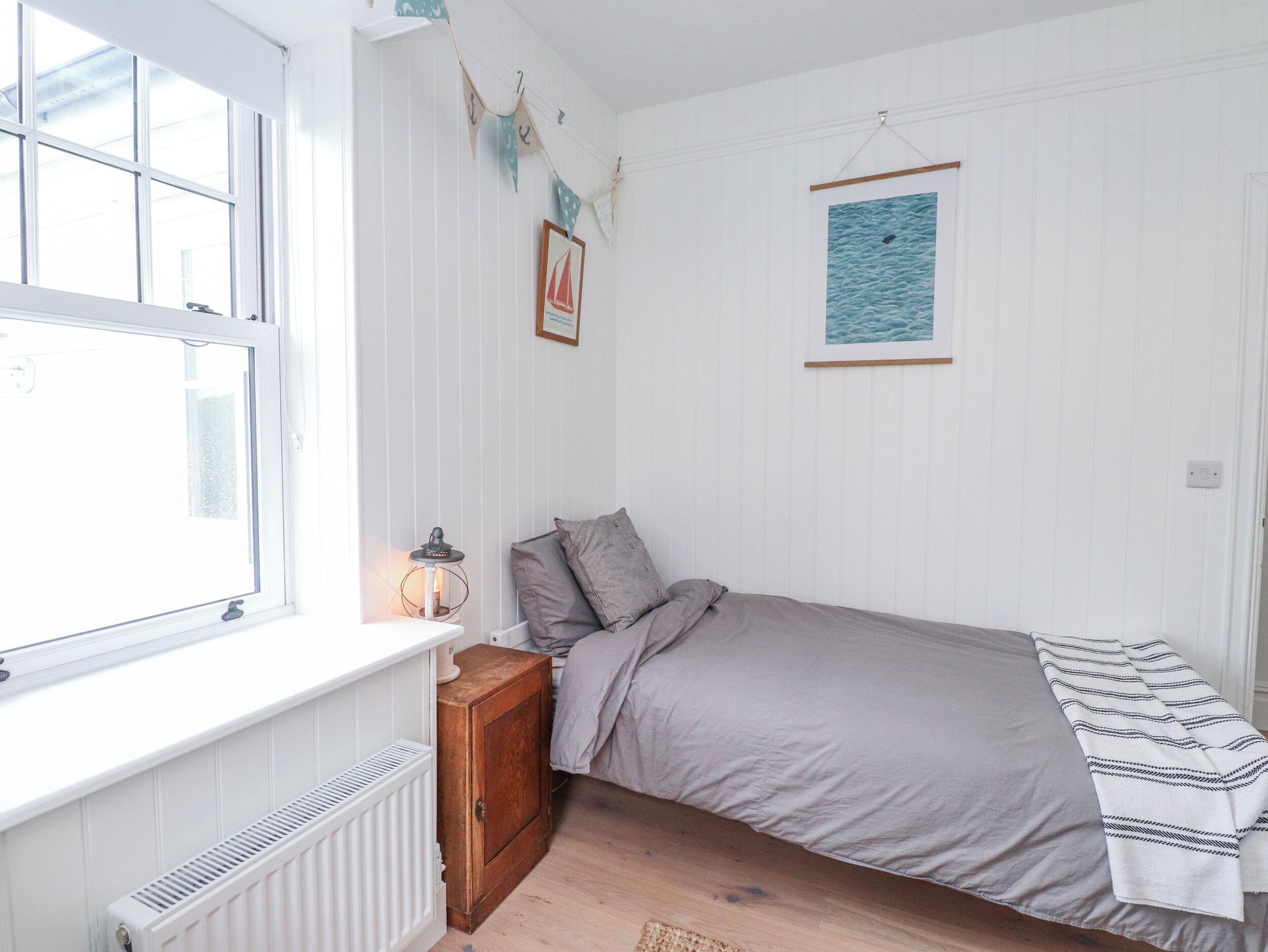 Un dormitorio con una cama individual, un radiador, un mueble de madera pequeño, un cuadro en la pared y una ventana con cortinas.