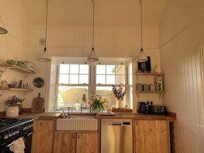 A kitchen with wooden cabinets, a stove, a sink, and a window with a view. - Arfryn (Amlwch)