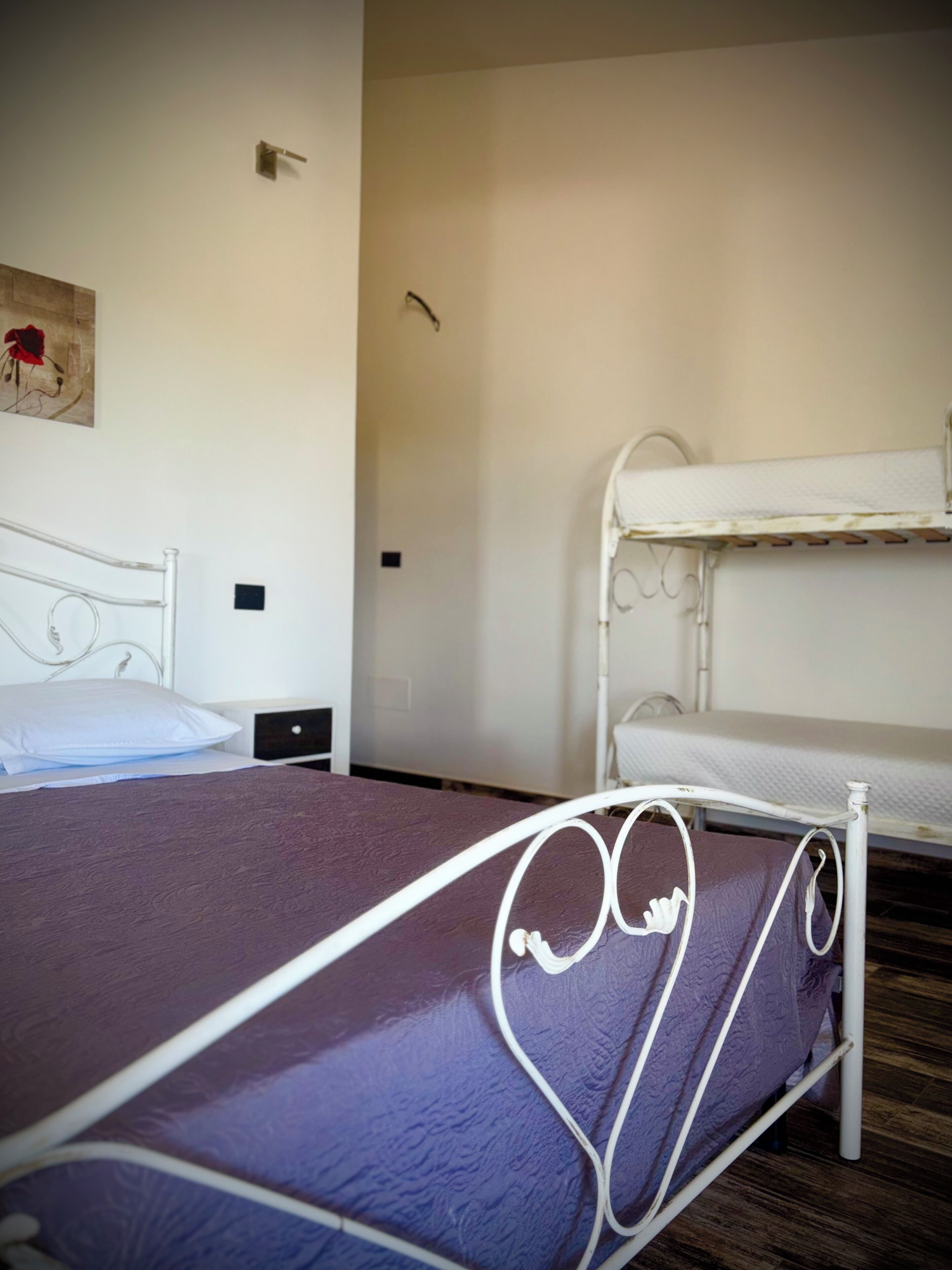 A bunk bed with a metal frame, a single bed with a white headboard, a small bedside table, and a wall-mounted light fixture.