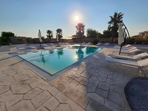 Una piscina rectangular con superficie de agua azul cristalina, rodeada de baldosas de piedra y sillas de descanso blancas.