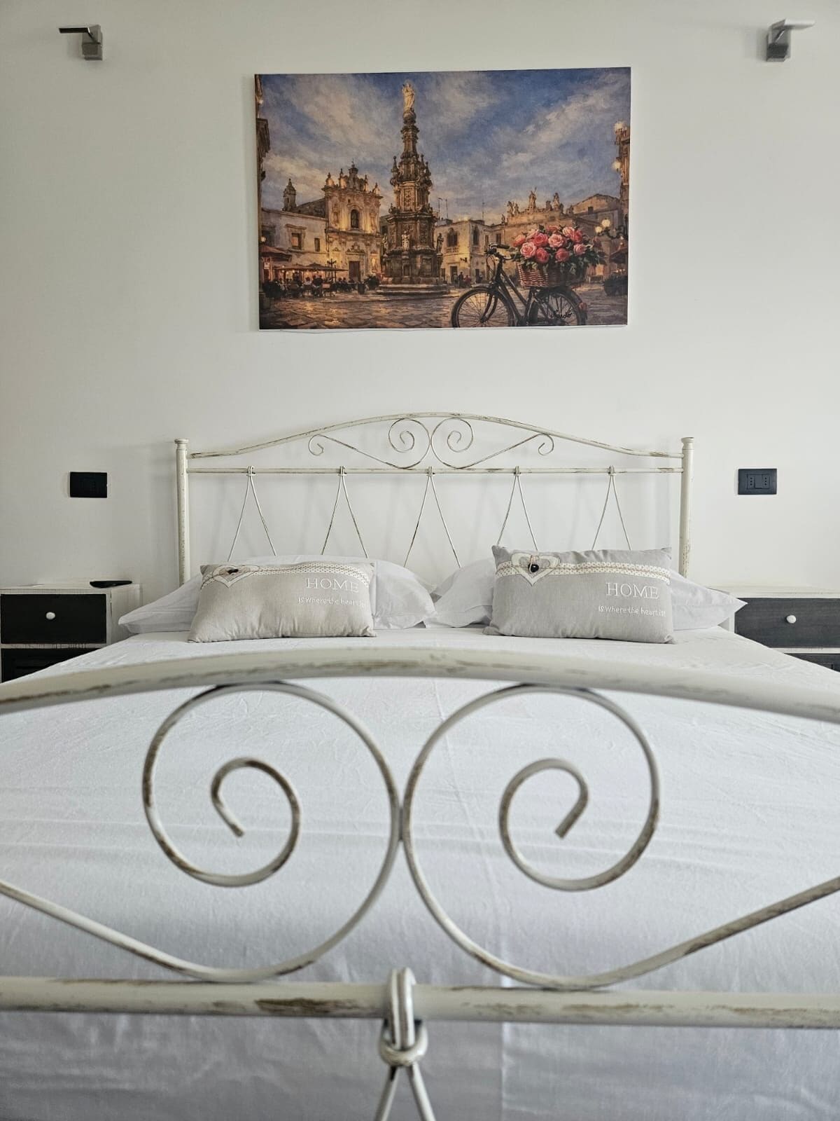 A bed with a metal headboard, white bedding, and two pillows with 'HOME' text. A painting of a building and flowers is on the wall.
