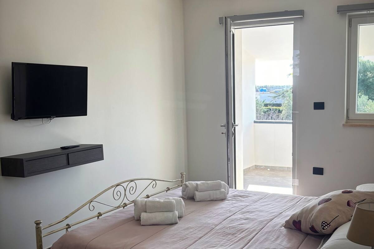 A bedroom with a bed, a TV mounted on the wall, a ceiling light, and a door leading to an outdoor area.