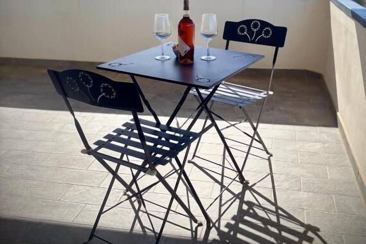 A small outdoor table with two chairs, a bottle, and two glasses on a balcony with a view of trees and buildings.