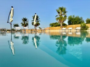 A clear swimming pool with lounge chairs and umbrellas, surrounded by palm trees and a wooden fence. - B&B Helios (Nardò)