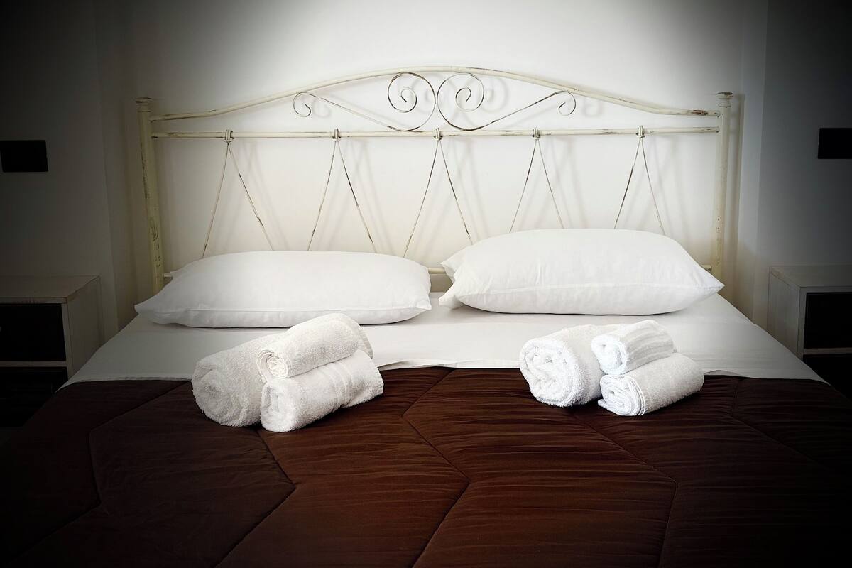 A bed with white linens, two pillows, and two rolled towels.