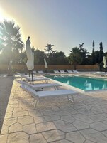 Zona de piscina con sillas de descanso y sombrillas, rodeada de árboles y un muro de piedra.
