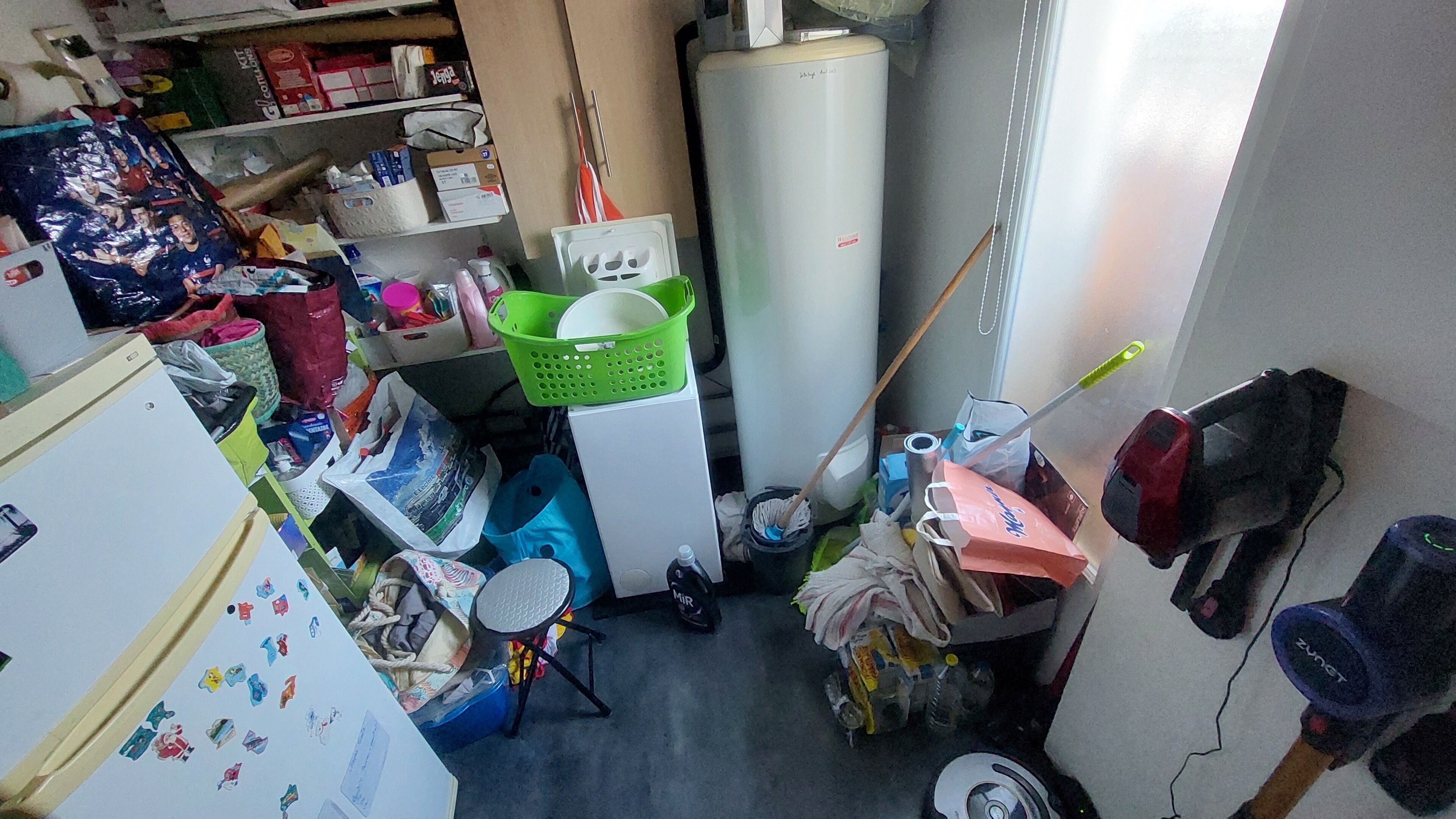 Une pièce encombrée où l’on trouve un réfrigérateur, une machine à laver, une serpillière, un balai et divers objets éparpillés.