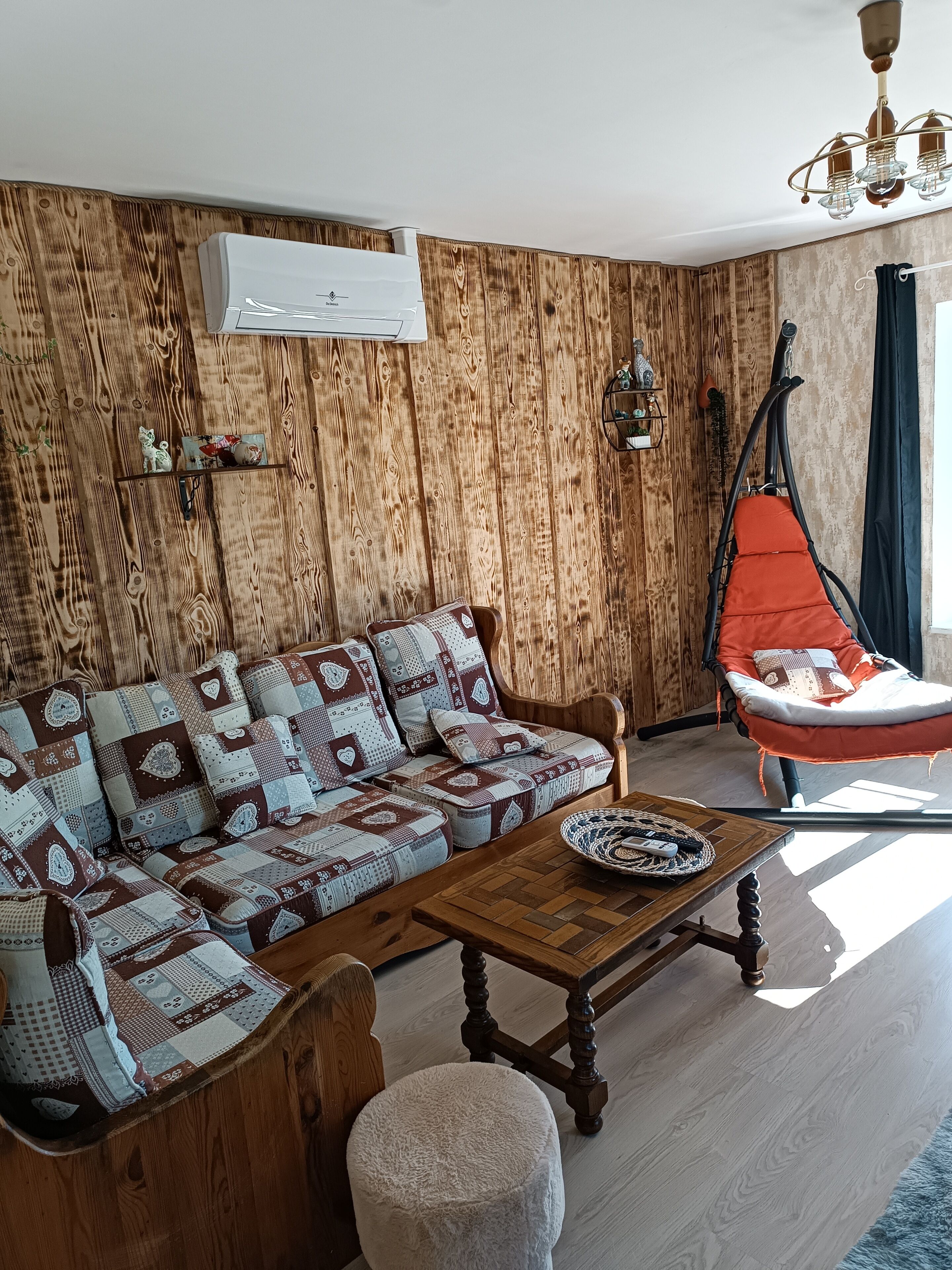 Un salon avec un lambris en bois, un canapé agrémenté de coussins à motifs, une table basse en bois, un fauteuil suspendu et un luminaire au plafond.