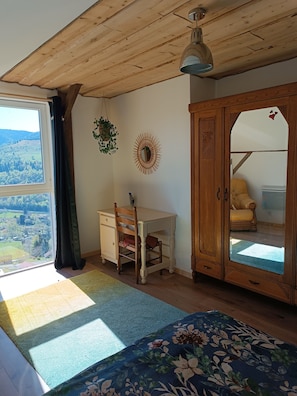 A room with a wooden ceiling, a desk with a chair, a wardrobe, and a view through a window.