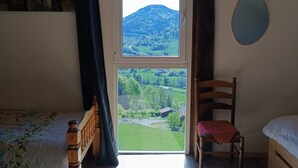 Une chambre avec un plafond en bois, une fenêtre donnant sur un paysage verdoyant, un lit recouvert d’une couvre-lit à motifs floraux, une chaise et une table de chevet.