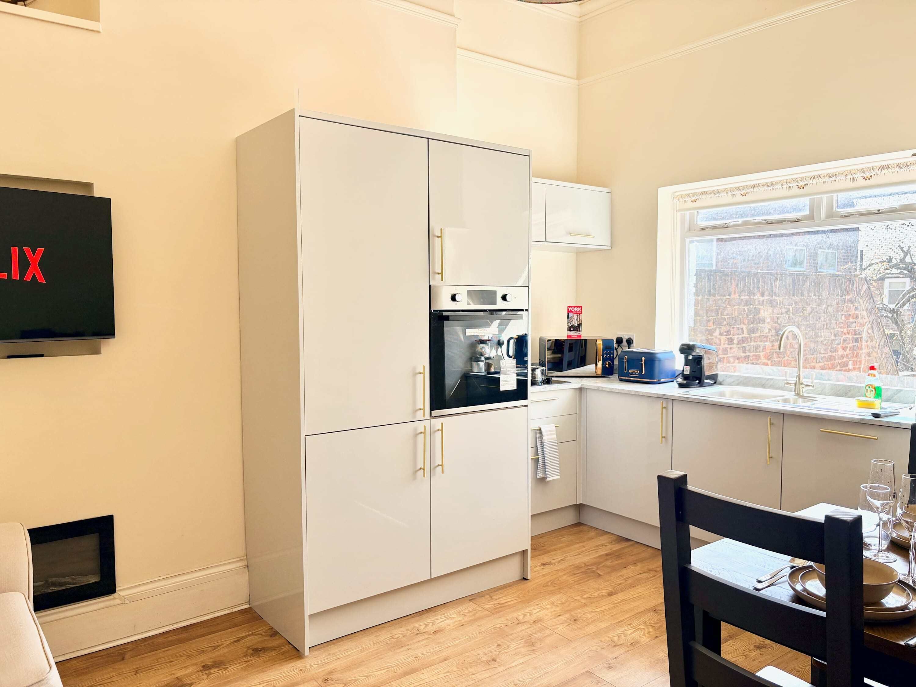 Una cucina moderna con mobili bianchi, forno integrato, microonde e tostapane. Si vede una finestra con vista su un edificio in mattoni.