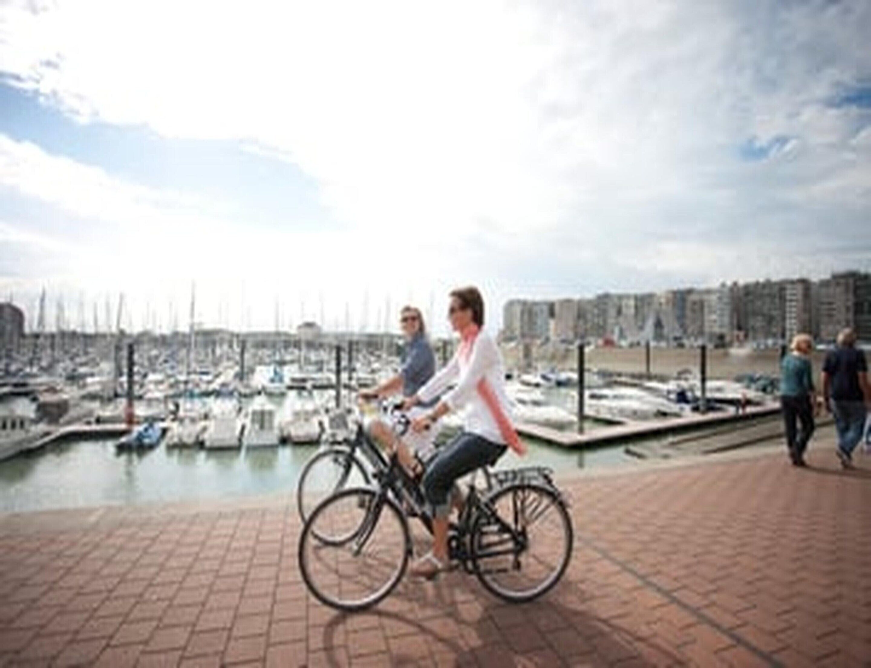 A marina with boats, a person on a bicycle, and a cityscape in the background.