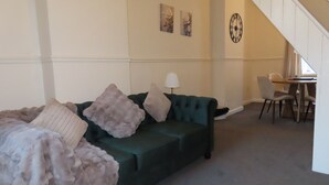 A living room with a green sofa, a table, and chairs. There is a staircase in the background.