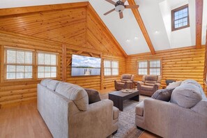 Interior de una cabaña de troncos con ventilador de techo, televisor grande y una zona de estar acogedora con sofás y sillones.