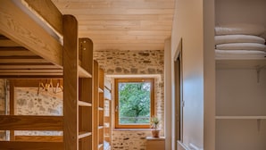 Ein Zimmer mit einer Holzleiter, einer Steinswand, einem Fenster mit Blick auf Bäume, einem kleinen Holztisch mit einer Pflanze und einem weißen Schrank.