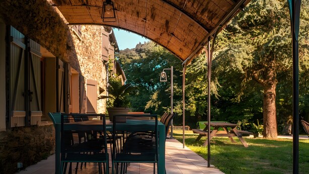 A covered patio area with a table and chairs, a stone wall, and a view of a wooded area. - Gîte De La Fontaine – Entire Property, Sleeps 13, Les Cammazes (Les Cammazes)