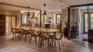 A spacious dining area with a large wooden table and chairs, a kitchen in the background, and a view of the outdoors through a window. - Gîte De La Fontaine – Entire Property, Sleeps 13, Les Cammazes (Les Cammazes)