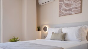 A neatly made bed with white linens, a wall-mounted air conditioner, a small potted plant, and a framed abstract artwork. - Aegean Essence Villa (Saronikos)