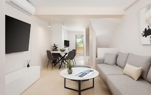 Una sala de estar moderna con un sofá gris, una mesa de centro redonda, un área de comedor con televisor y vistas a un balcón.