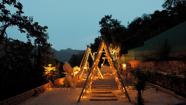 A nighttime outdoor area with a wooden structure illuminated by string lights, surrounded by trees and a stone wall. - SWAR Kasauli - Suites Cuisine Cave Club (Kasauli)