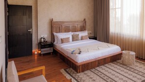 A bedroom with a bed, a chair, a nightstand, a ceiling fan, and a window with curtains. - SWAR Kasauli - Suites Cuisine Cave Club (Kasauli)