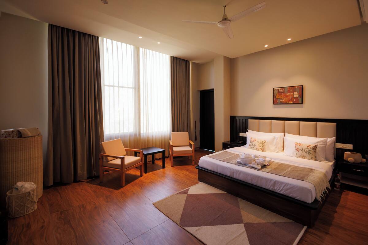 Quarto de hotel com uma cama grande, duas cadeiras, uma mesinha, uma lixeira e um ventilador de teto.
