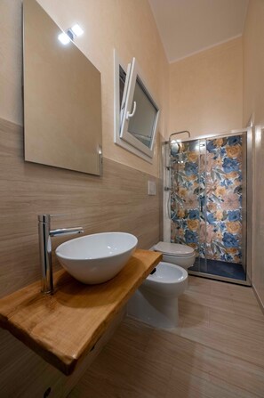 A bathroom with a wooden vanity, a white sink, a toilet, and a shower with floral tiles. - Suite degli Ulivi - Teresita Guest House (Gallipoli)