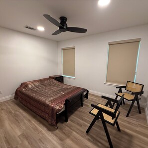 Un dormitorio con una cama, un ventilador de techo, una ventana con persianas, una silla y una mesita de noche.