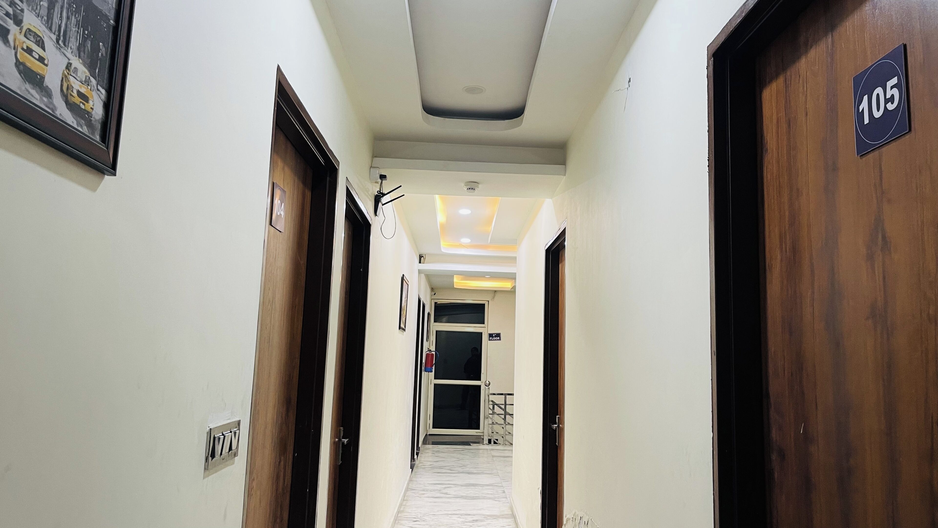 A hotel hallway with wooden doors, numbered signs, a fire extinguisher, and a ceiling with recessed lighting.