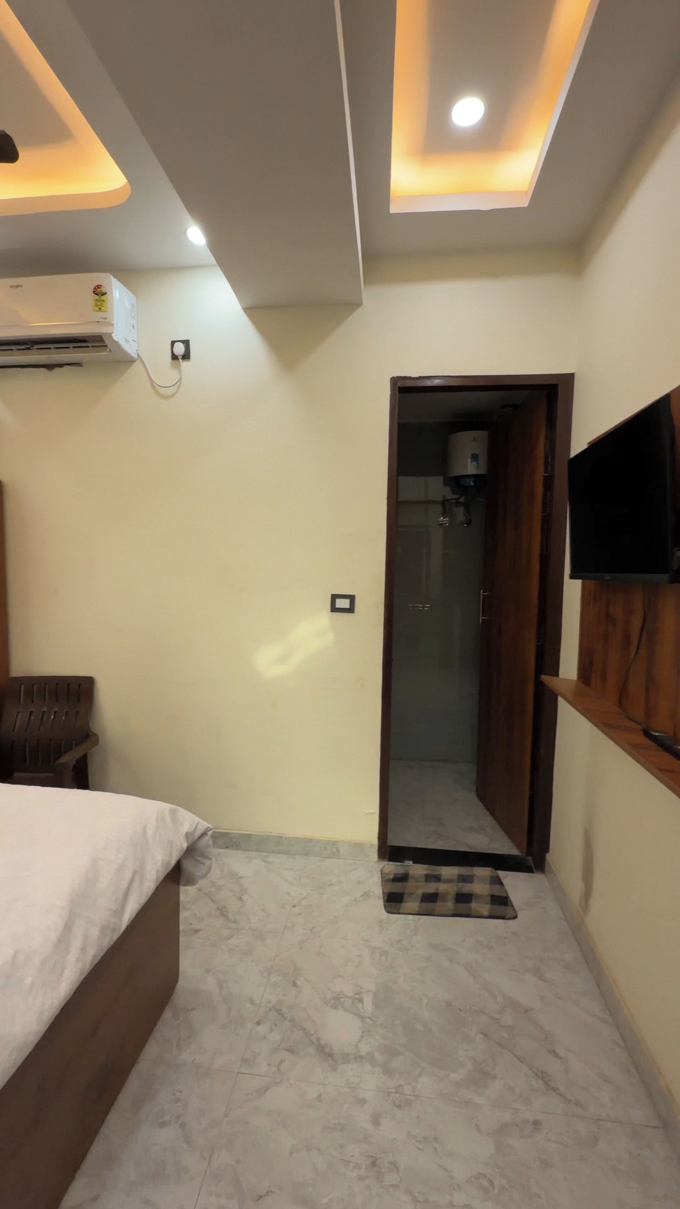 Une chambre à coucher comprenant un lit, un climatiseur, un téléviseur fixé au mur et une porte donnant accès à une salle de bain.