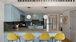 Una cocina con armarios azules, una isla central con taburetes, electrodomésticos de acero inoxidable y una lámpara colgante.