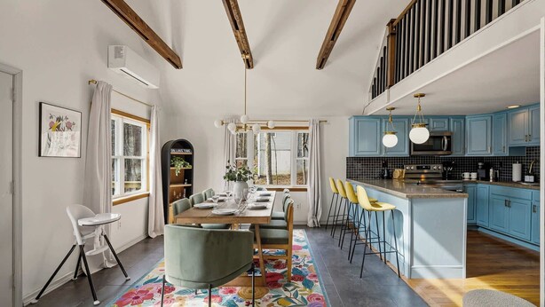 Un comedor con una mesa preparada para cuatro, techo alto con vigas a la vista, y una cocina con armarios azules y una isla central.