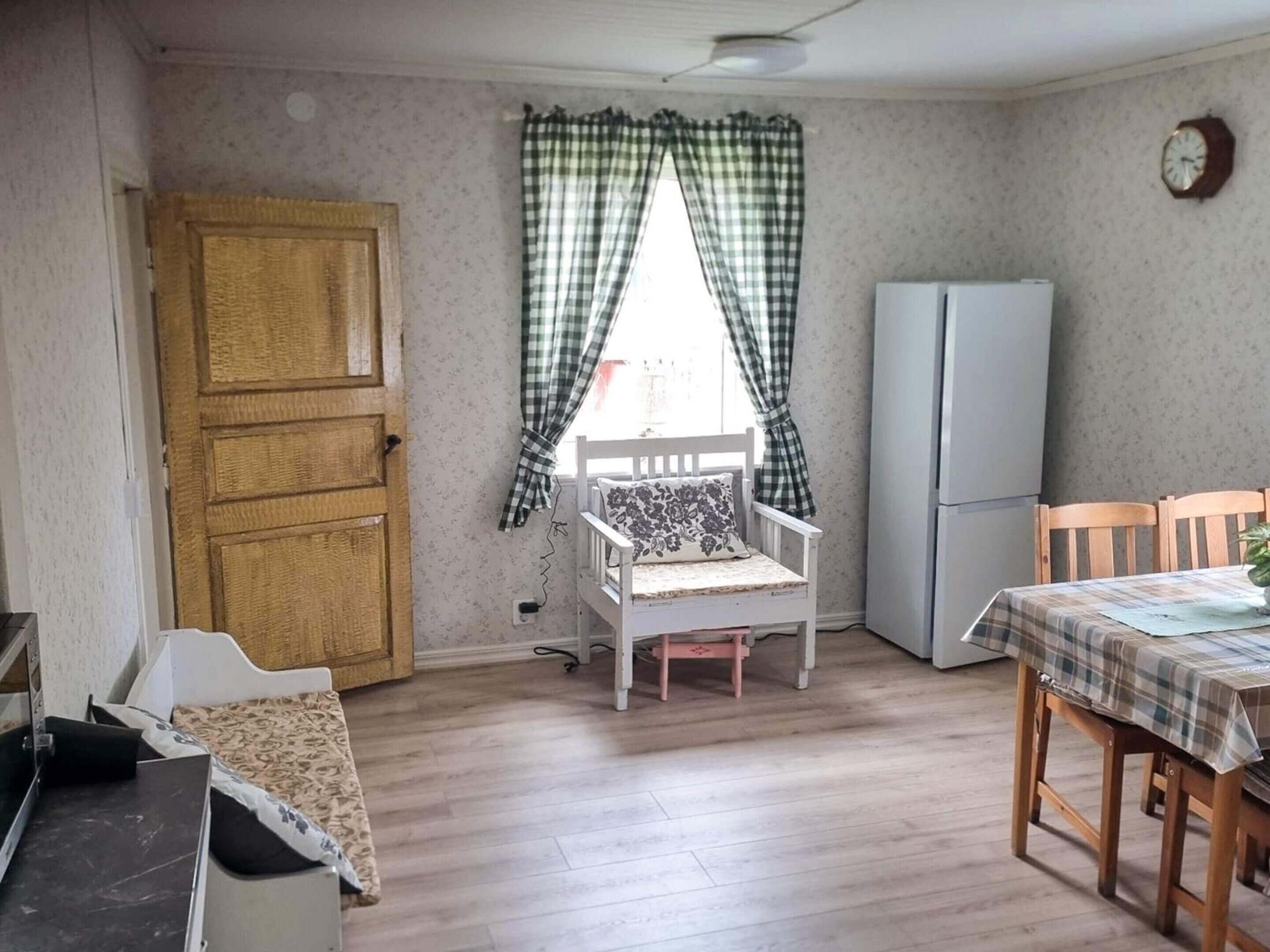 A room with a wooden door, a white bench, a table with a plaid tablecloth, a refrigerator, and a window with checkered curtains.