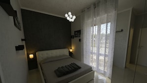 A bedroom with a bed, bedside lamps, a chandelier, and a view through a window with curtains.