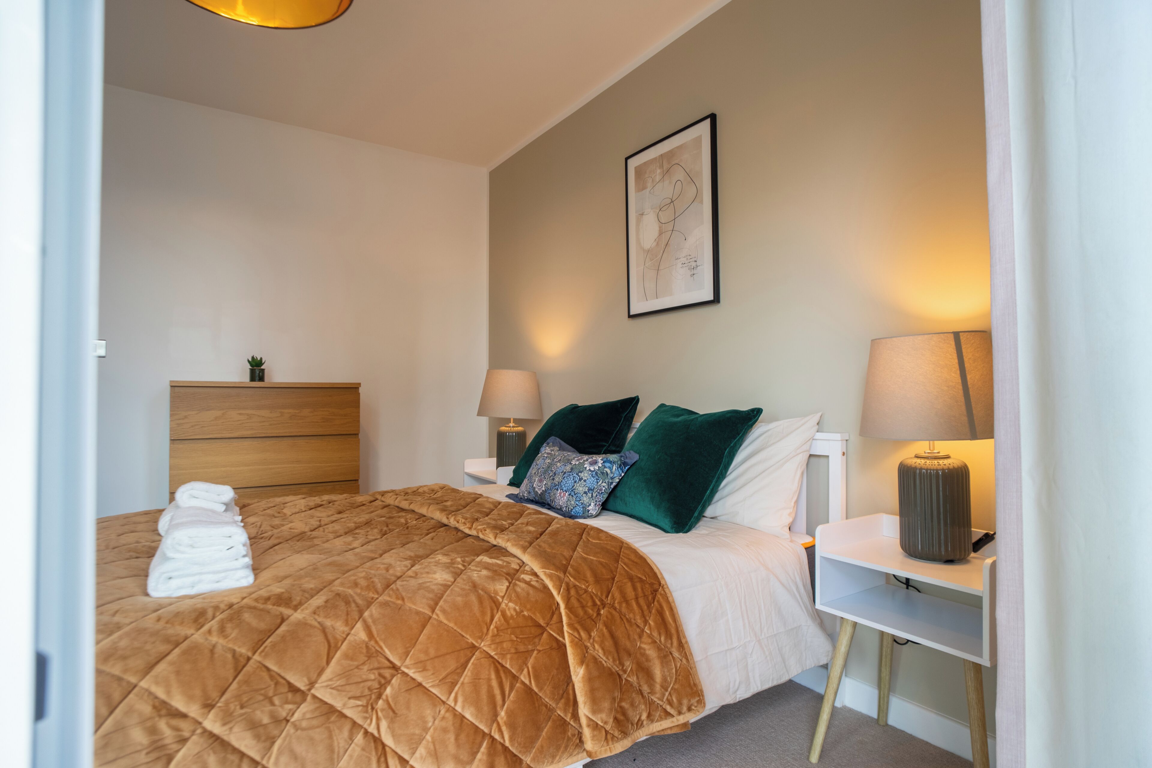 Hotellihuone, jossa on sänky, yöpöydät, lamppu, seinätaideteos ja pieni kasvi.