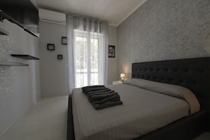 A bedroom with a bed, a nightstand, a lamp, a window with curtains, and a wall-mounted air conditioning unit. - Double Room 1 with Wi-Fi and Air Conditioning (Baia Verde)