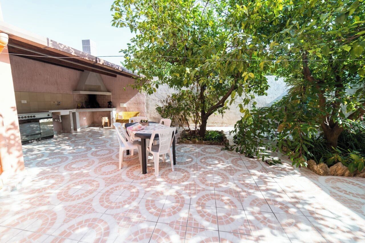 Une terrasse extérieure avec un sol carrelé, une table entourée de chaises, un barbecue et un arbre qui offre de l’ombre.
