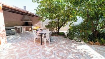 Une terrasse extérieure avec un sol carrelé, une table entourée de chaises, un barbecue et un arbre qui offre de l’ombre.