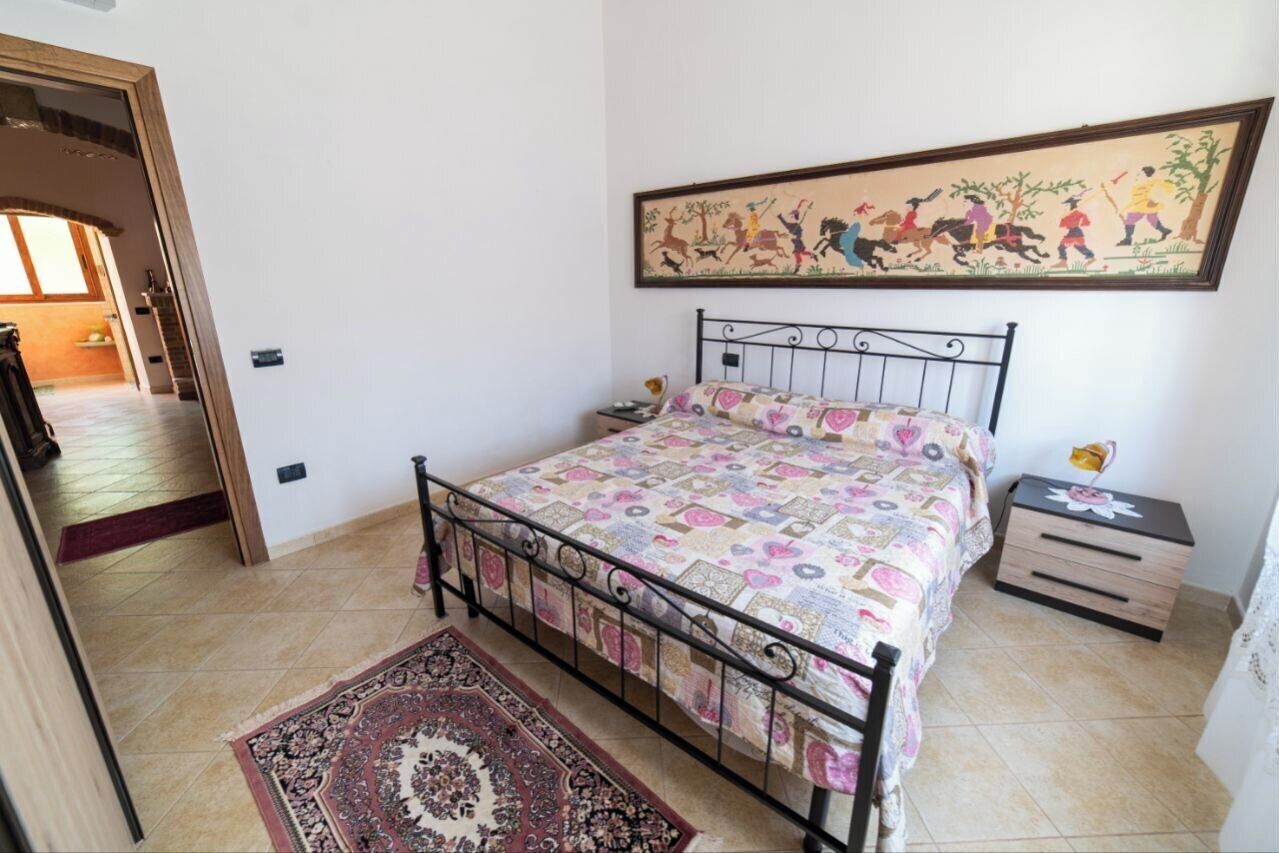 A bedroom with a bed, a nightstand, a wall-mounted painting, and a door leading to another room.