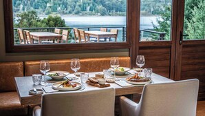 Un área de comedor con una mesa lista para dos personas, vista a un lago y un balcón con sillas.