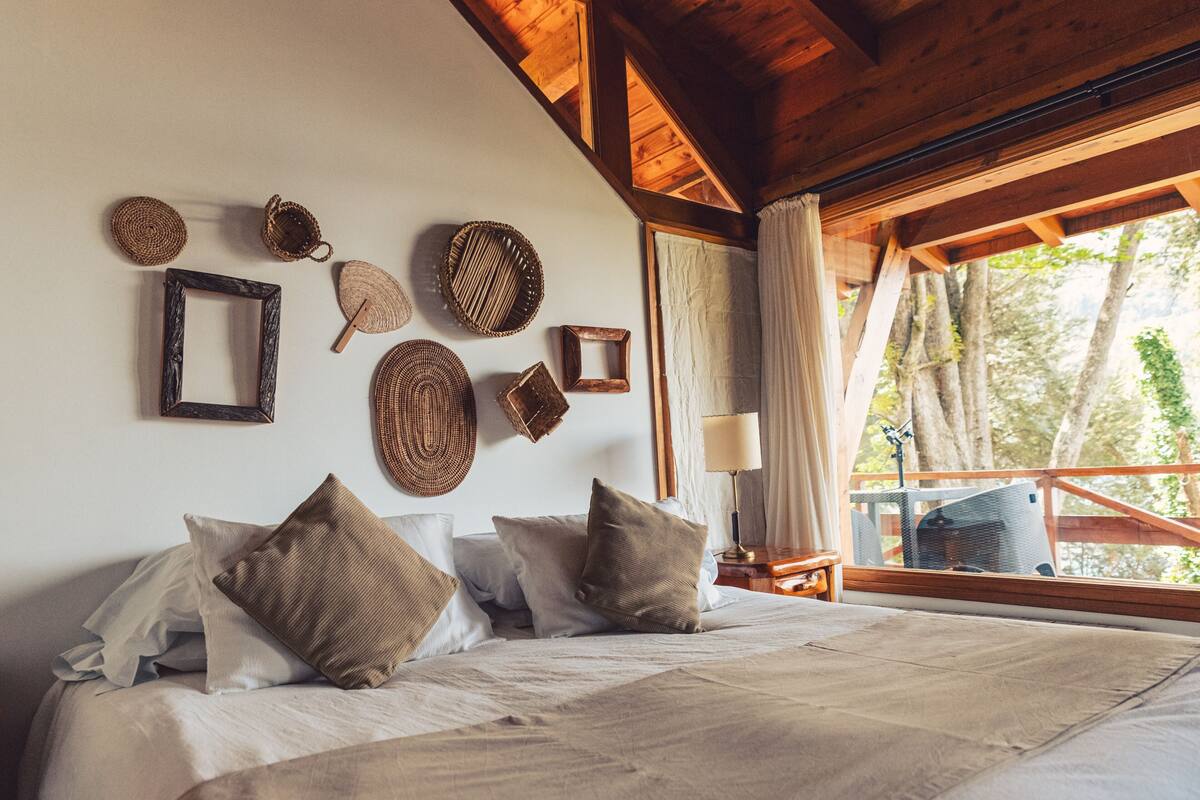 Un dormitorio con una cama grande, varios almohadones, techo de madera y ventana con vista a árboles.