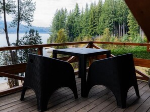 Una terraza de madera con dos sillas de mimbre y una mesa pequeña, con vistas a un lago y un bosque.