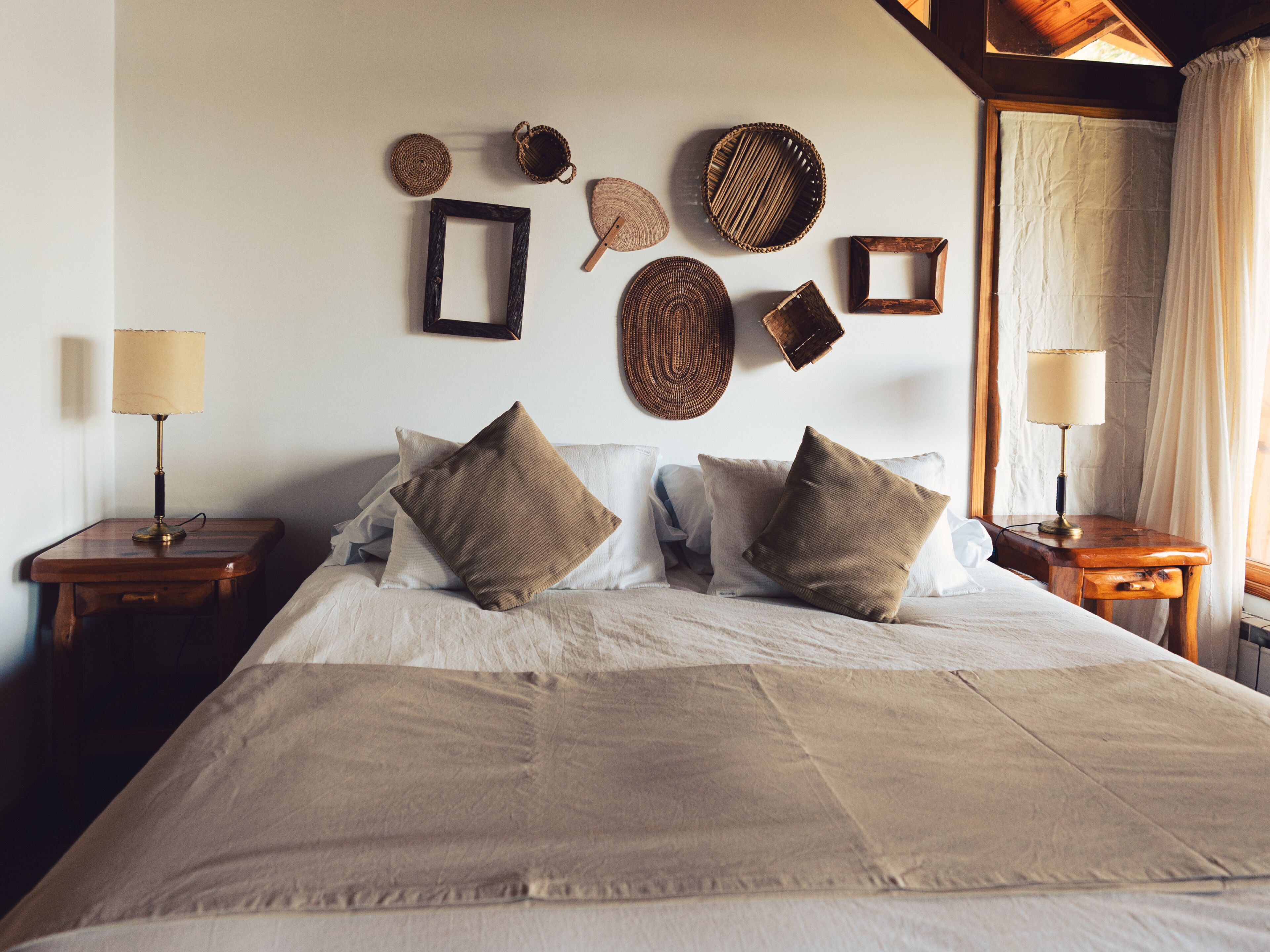 A bed with a beige comforter and pillows, two bedside tables with lamps, and wall decor including woven baskets and a framed picture.