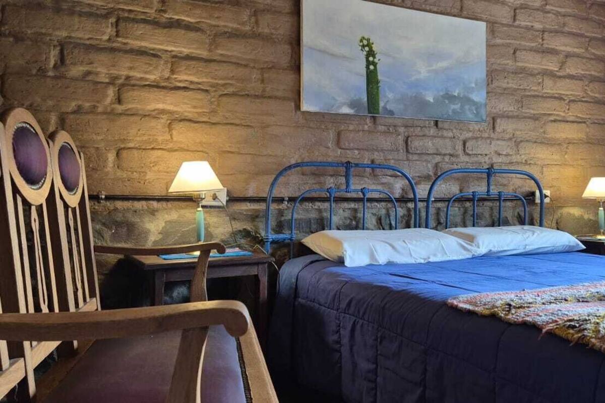 A room with a stone wall, a bed with a blue bedspread, two nightstands with lamps, and a wooden chair.
